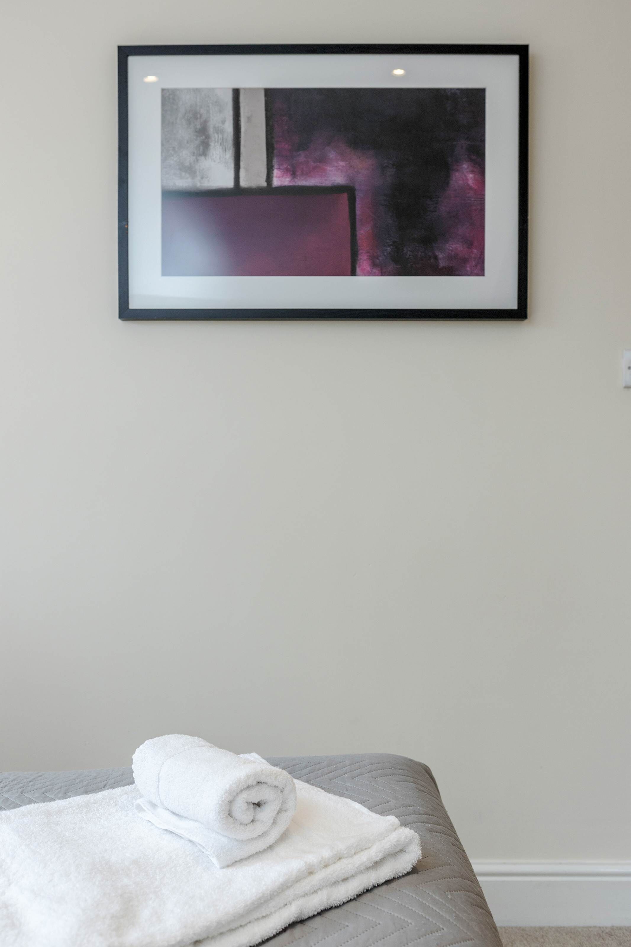 Un cuadro abstracto enmarcado en una pared, una toalla blanca doblada con cuidado sobre una cama gris.