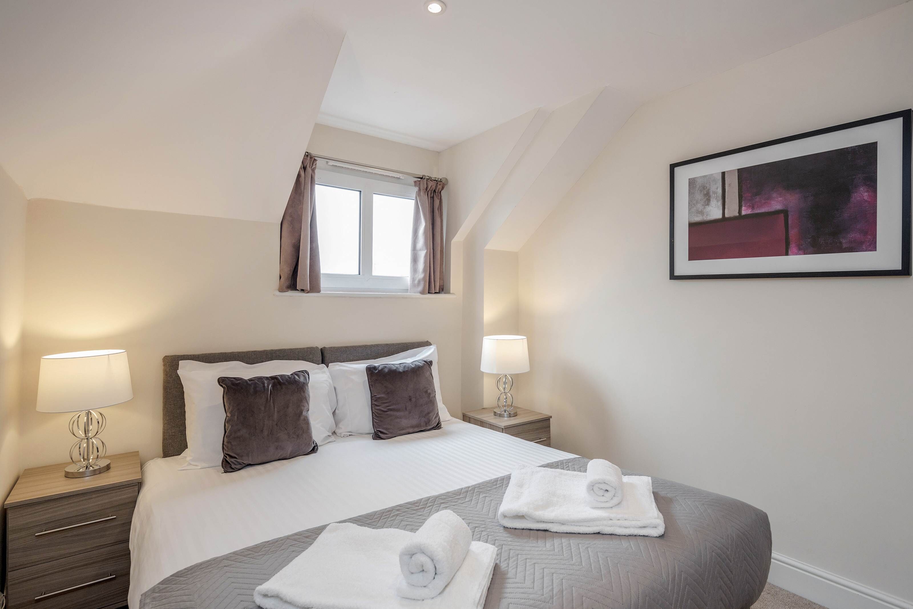 A neatly arranged hotel room with a large bed, two bedside tables with lamps, a window with curtains, and a framed artwork on the wall.