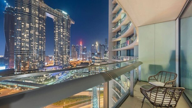 Un immeuble de grande hauteur doté d’un balcon où se trouvent deux fauteuils en osier et une balustrade en verre, offrant une vue sur le paysage urbain illuminé la nuit.