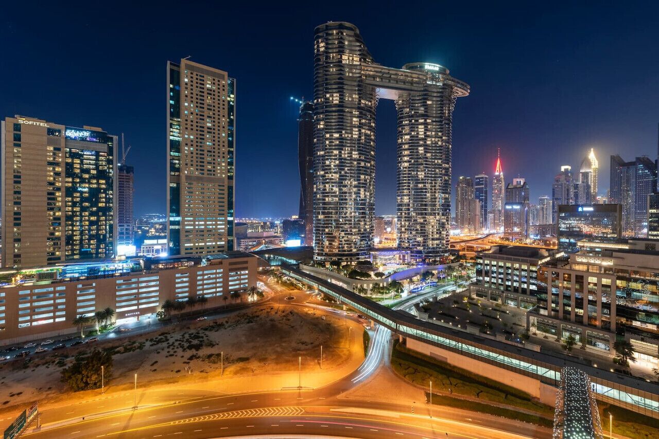 A cityscape at night with modern high-rise buildings, including a distinctive curved structure, and a well-lit highway with visible traffic.