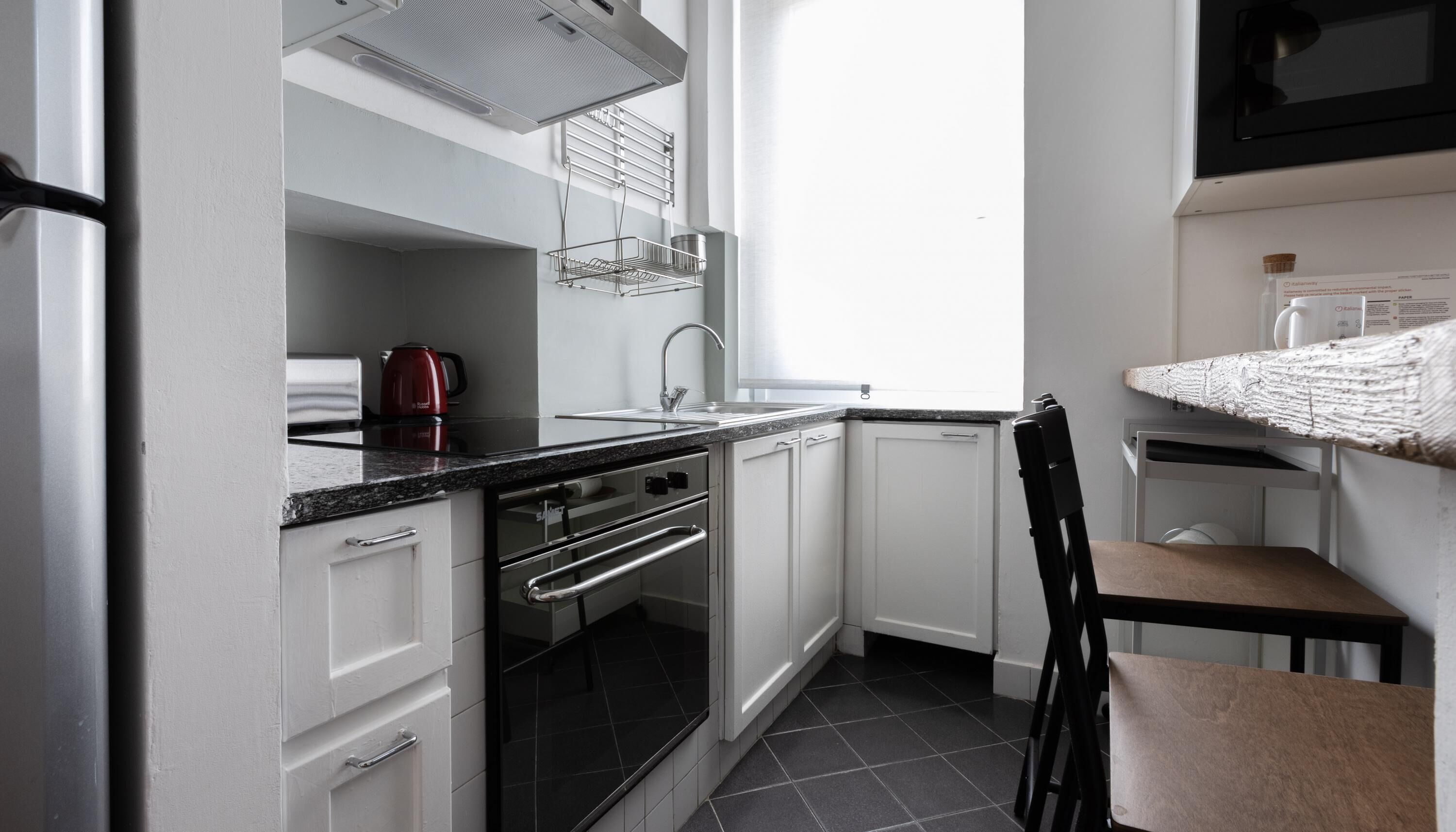 A compact kitchen with white cabinets, a black countertop, a stainless steel sink, and a red kettle.