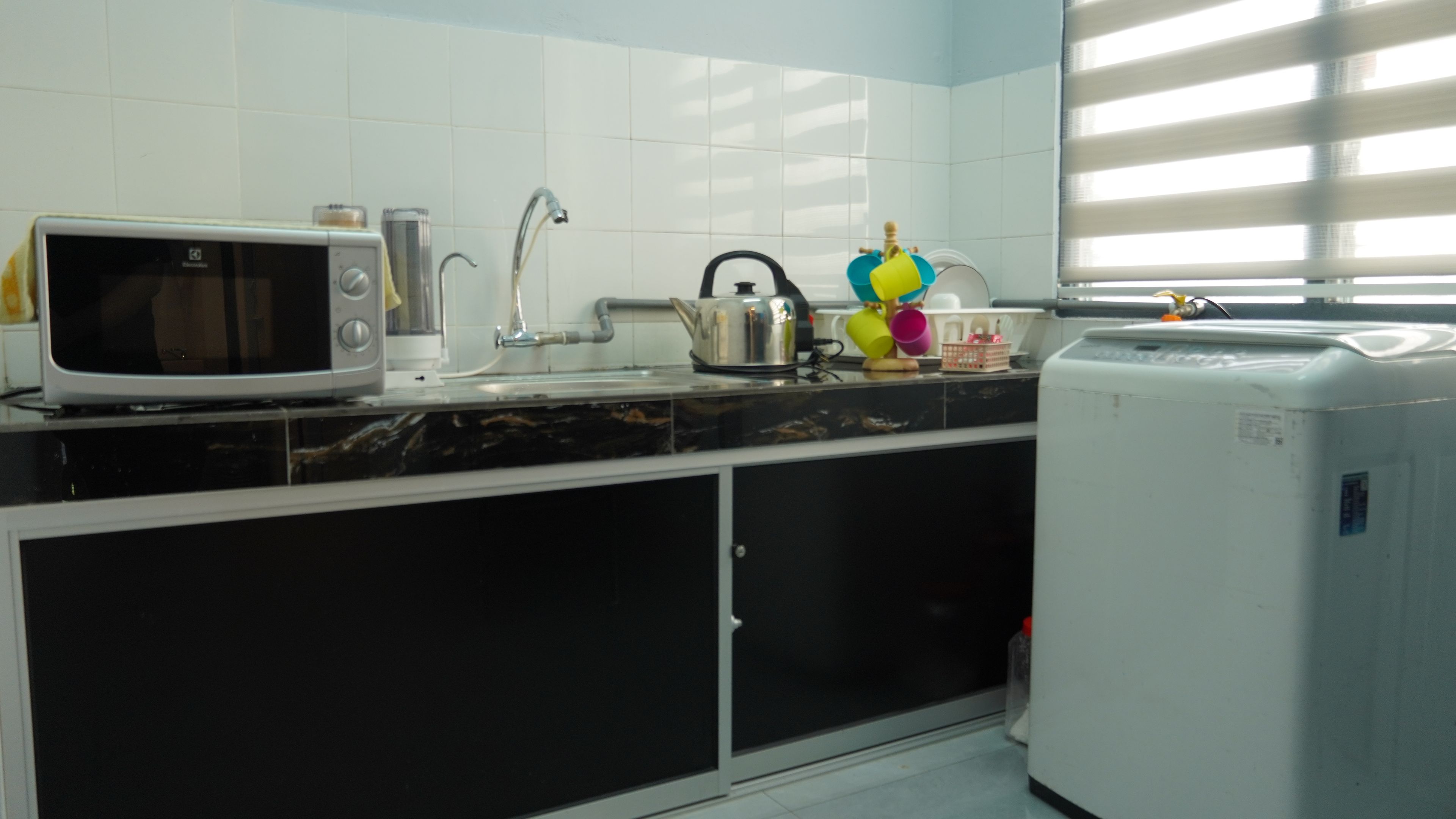 A kitchen with a microwave, kettle, sink, and a washing machine.