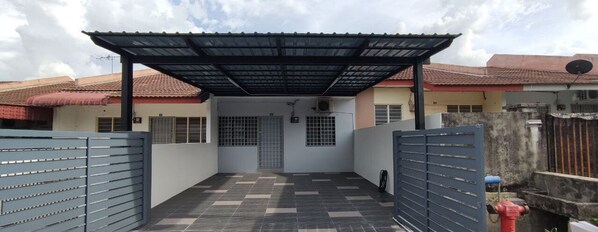 A covered outdoor area with a tiled floor, metal gates, and a building with a red roof in the background. - Margosa A Stay Botani Ipoh Homestay 3 BR (Ipoh)