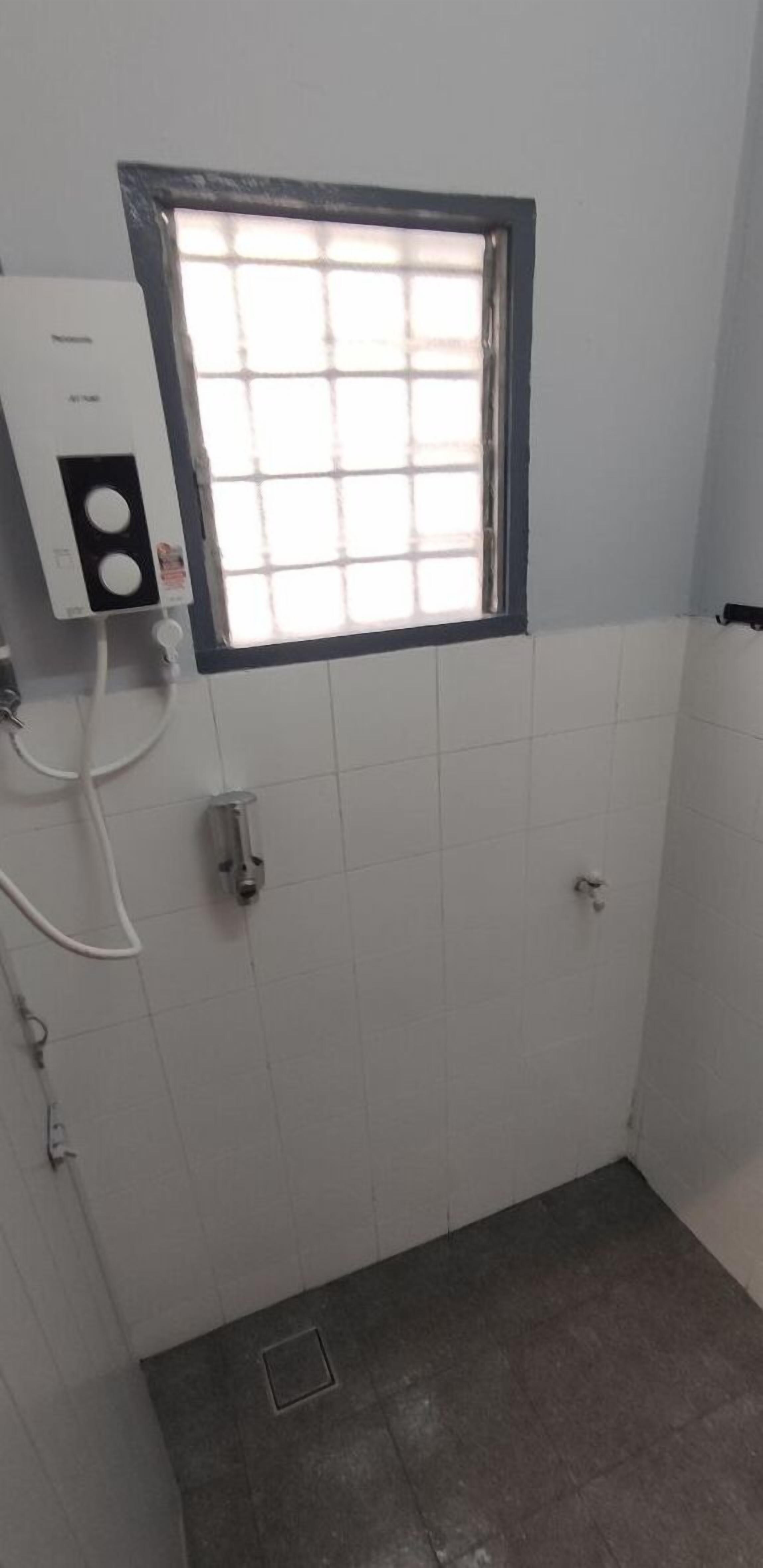 Une petite salle de bain aux murs recouverts de carreaux blancs, une fenêtre à motifs géométriques, une boîte électrique blanche et un robinet de douche.