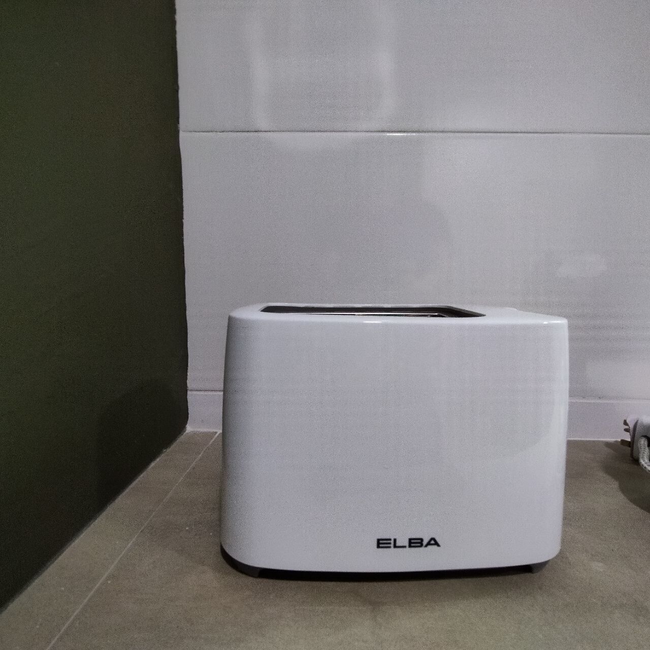 A white ELBA portable air purifier in a corner of a room with a tiled floor and a plain wall.