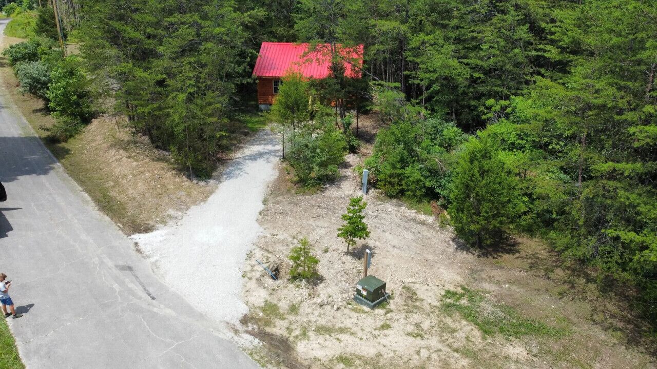 Uma cabana pequena com telhado vermelho, rodeada por uma floresta densa, com uma trilha de terra que leva até ela e uma caixa de serviços por perto.