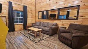 Ein gemütliches Blockhaus-Interieur mit Holzboden, einer Couch, einem Sessel, einem kleinen Tisch und einem Fenster mit Vorhängen.