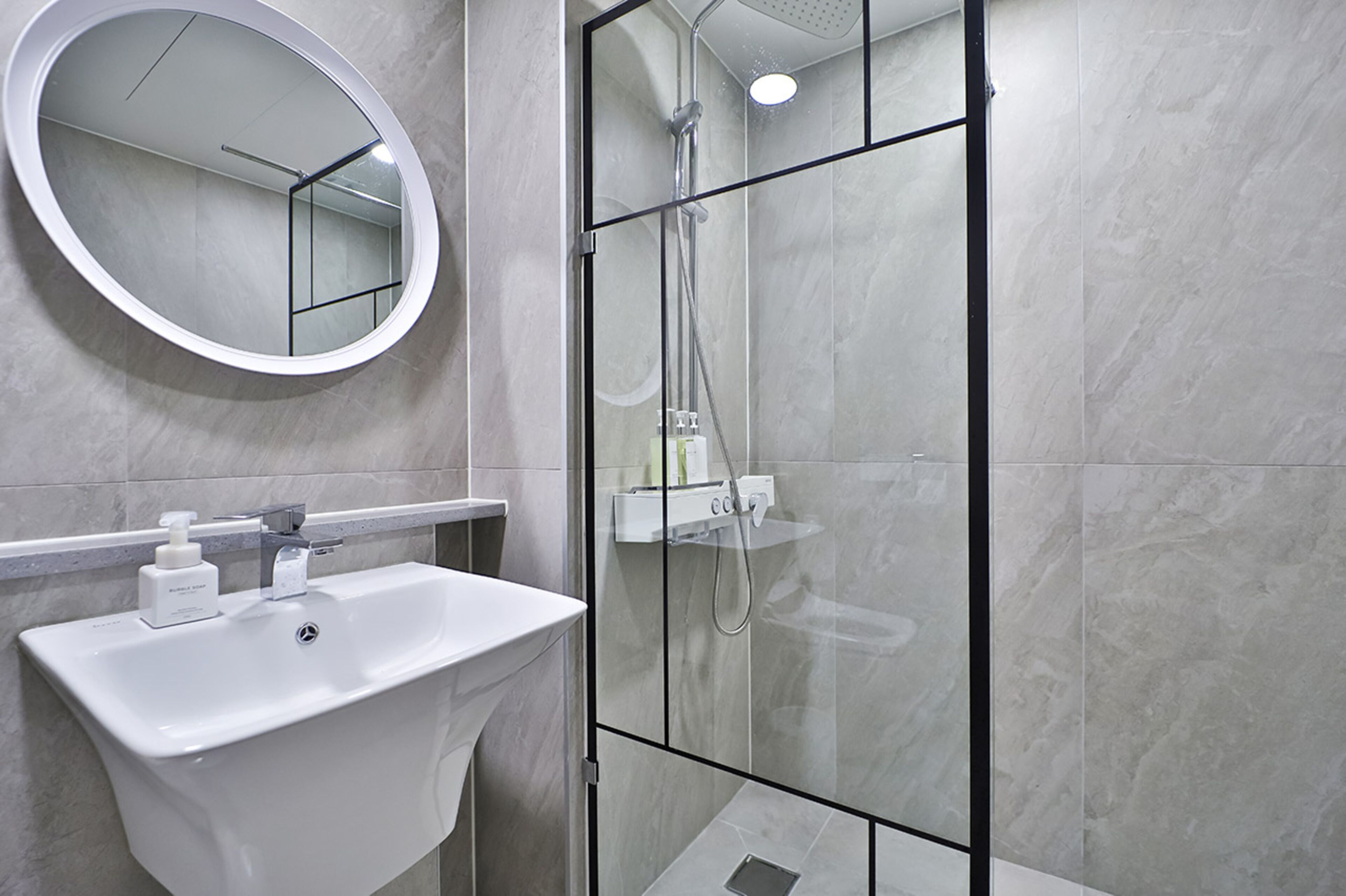 Un baño moderno con un lavamanos blanco, un espejo redondo y una ducha con mampara de vidrio.