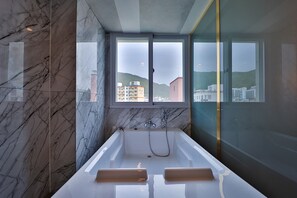 A modern bathroom with a bathtub, marble walls, and a large window offering a city view. - Gimhae Eobang-dong City Hotel (Gimhae)