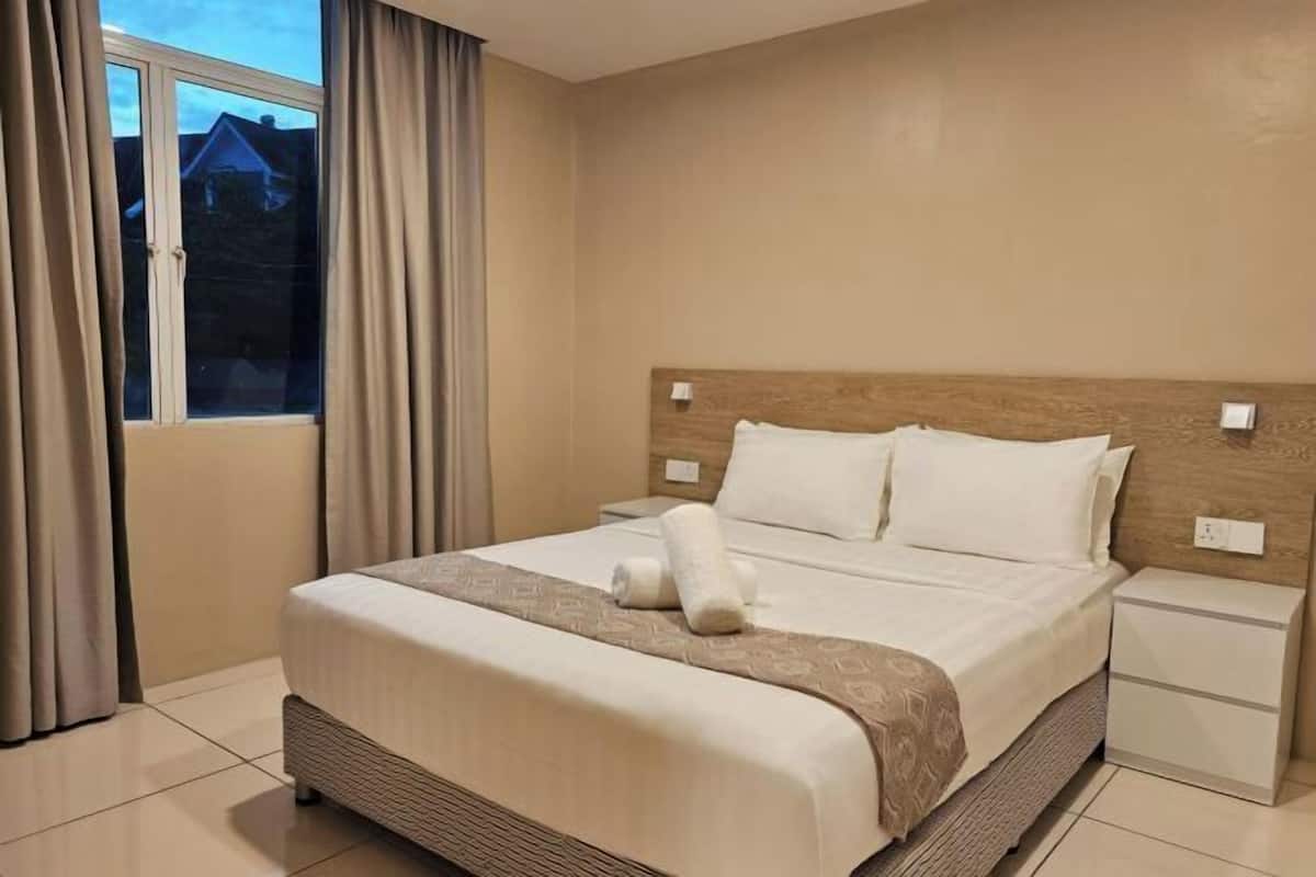 Una habitación de hotel con una cama grande, una mesita de noche, una ventana con cortinas y vistas a casas desde afuera.