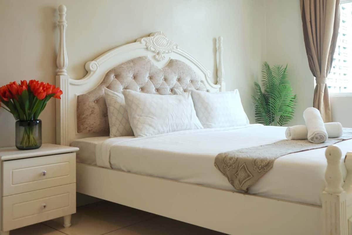 Una cama con dosel blanco con cabecera capitonada, una mesita de noche con un jarrón de flores rojas y una ventana con cortinas.