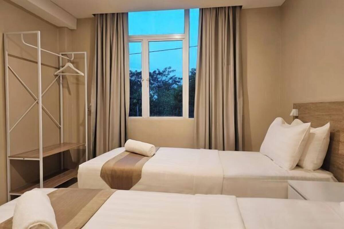 Habitación de hotel con dos camas, una ventana con cortinas, un perchero y un cabecero de madera.