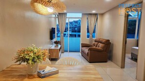 A modern living room with a sofa, a dining table with a vase of flowers, and a view of a building through the window. - Triima Inn Dayang Bay by Perfect Host (Langkawi)