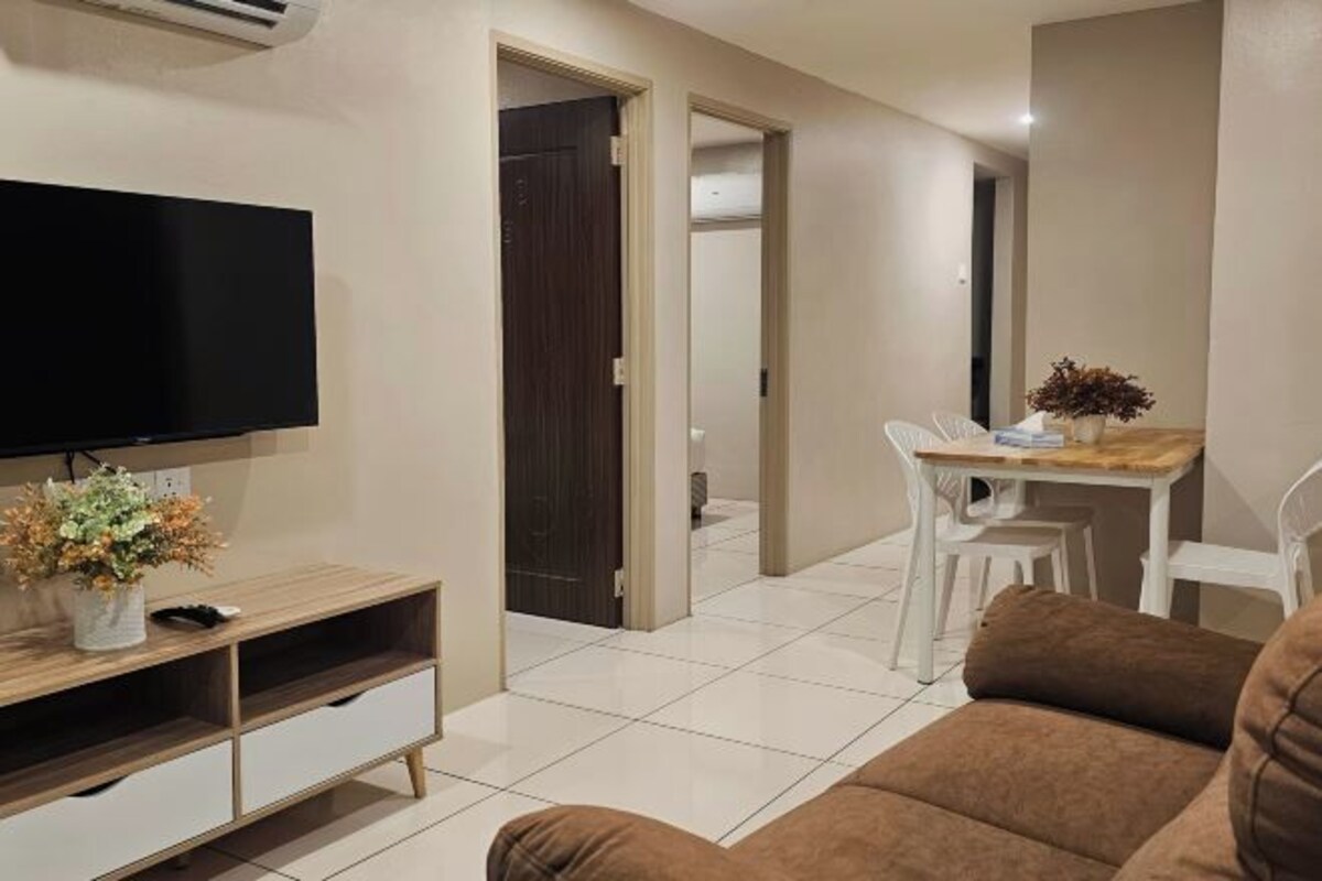 A living room with a flat-screen TV, a wooden entertainment unit, a brown sofa, a dining area with a table and chairs, and a hallway leading to another room.