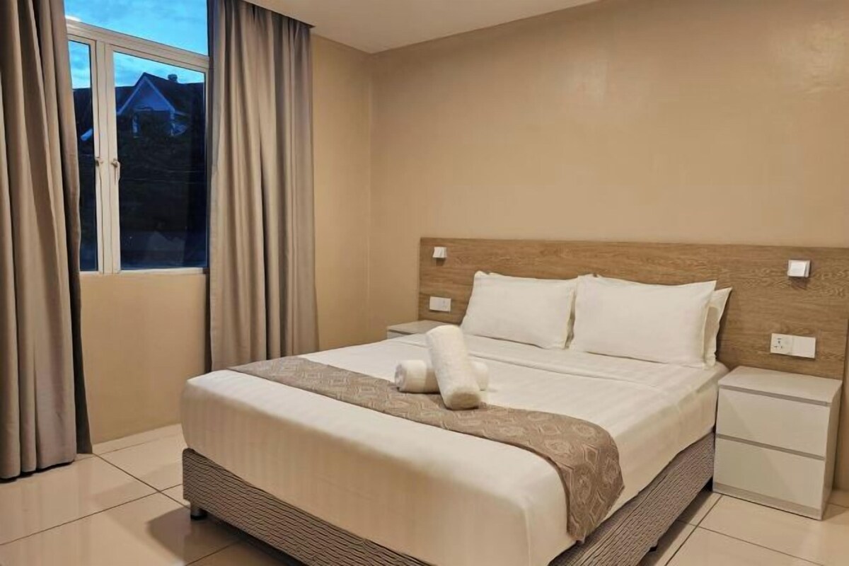 A hotel room with a large bed, a nightstand, a window with curtains, and a view of houses outside.