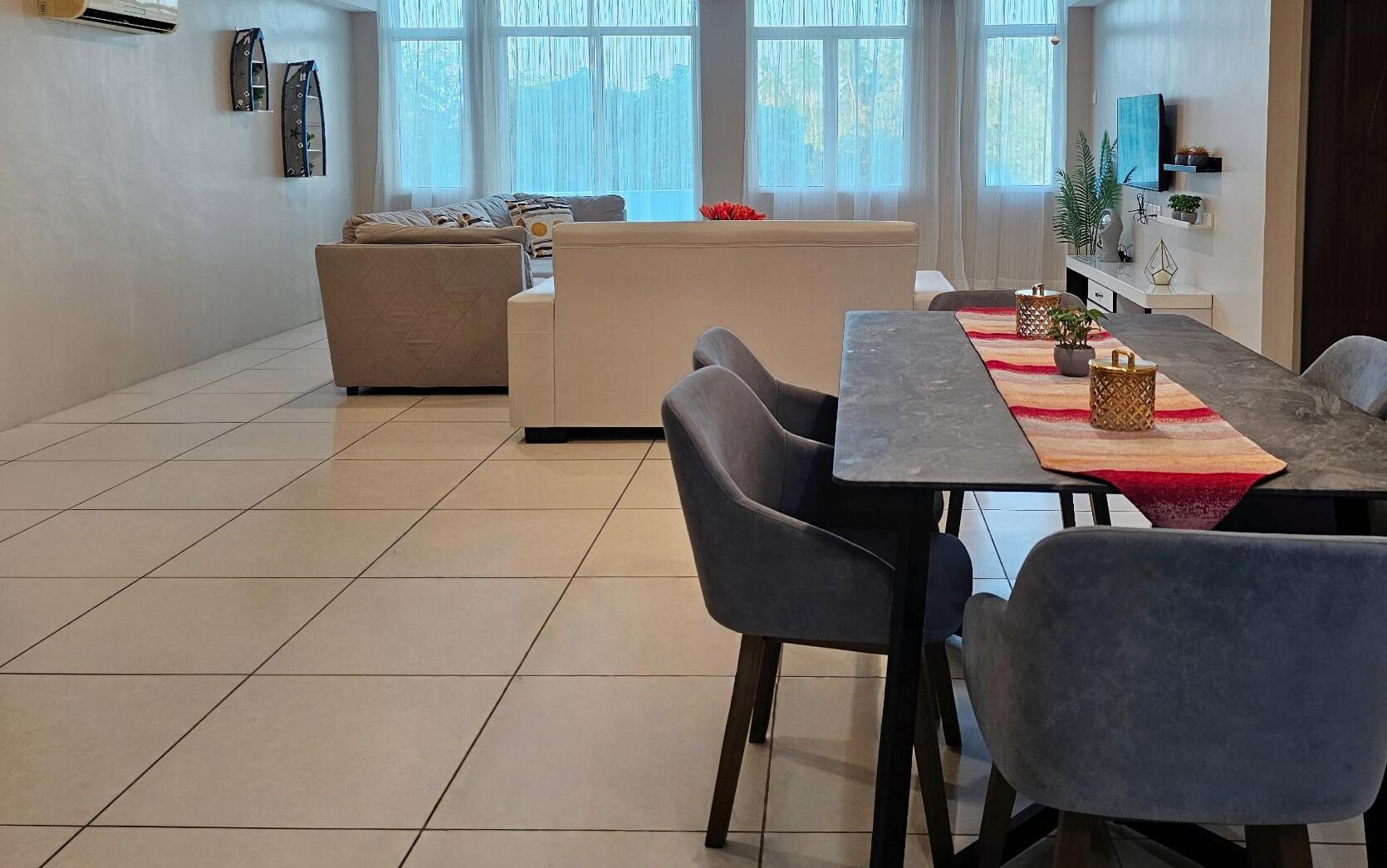 A modern living room with a sofa, dining table, and chairs. There are large windows with curtains, a flat-screen TV, and a small plant on the wall.