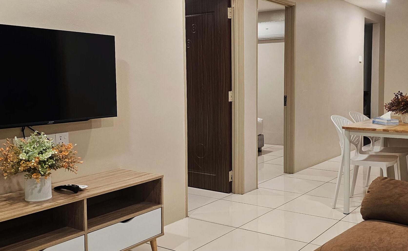 A modern living room with a flat-screen TV, a wooden entertainment unit, a dining area with a table and chairs, and a hallway leading to a bedroom.