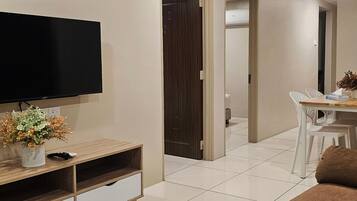 A modern living room with a flat-screen TV, a wooden entertainment unit, a dining area with a table and chairs, and a hallway leading to a bedroom.