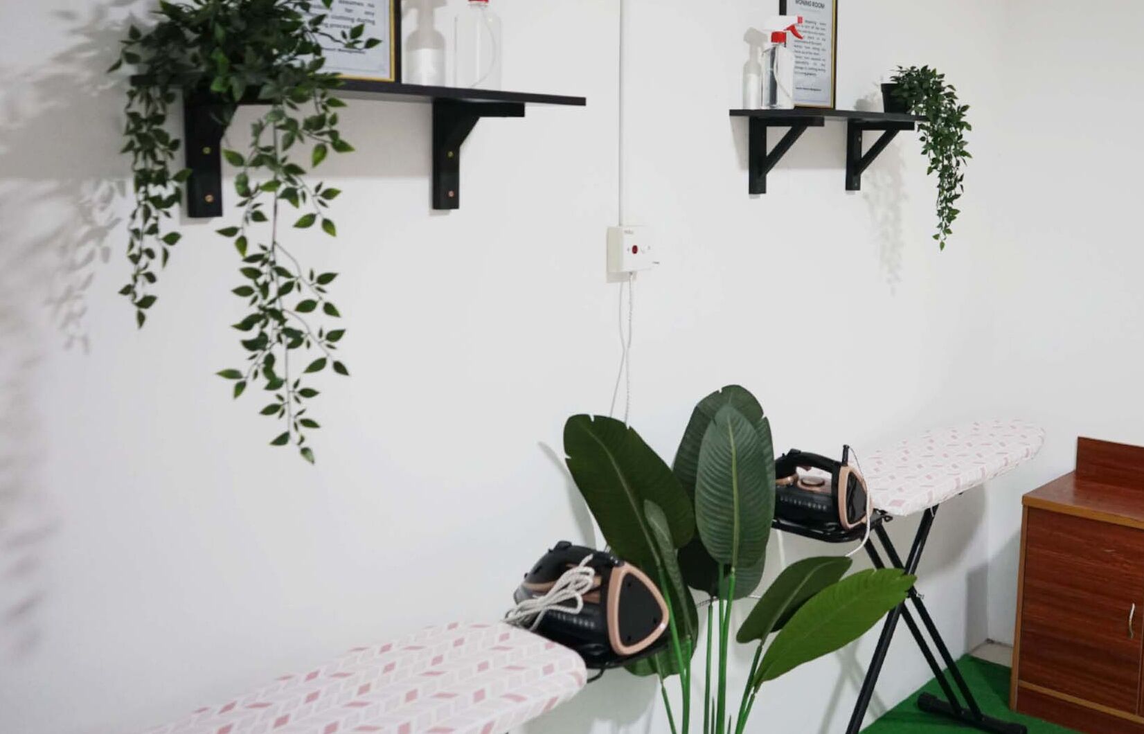 A room with a wooden cabinet, a white ironing board with a patterned cloth, and two black shelves with plants and cleaning supplies.