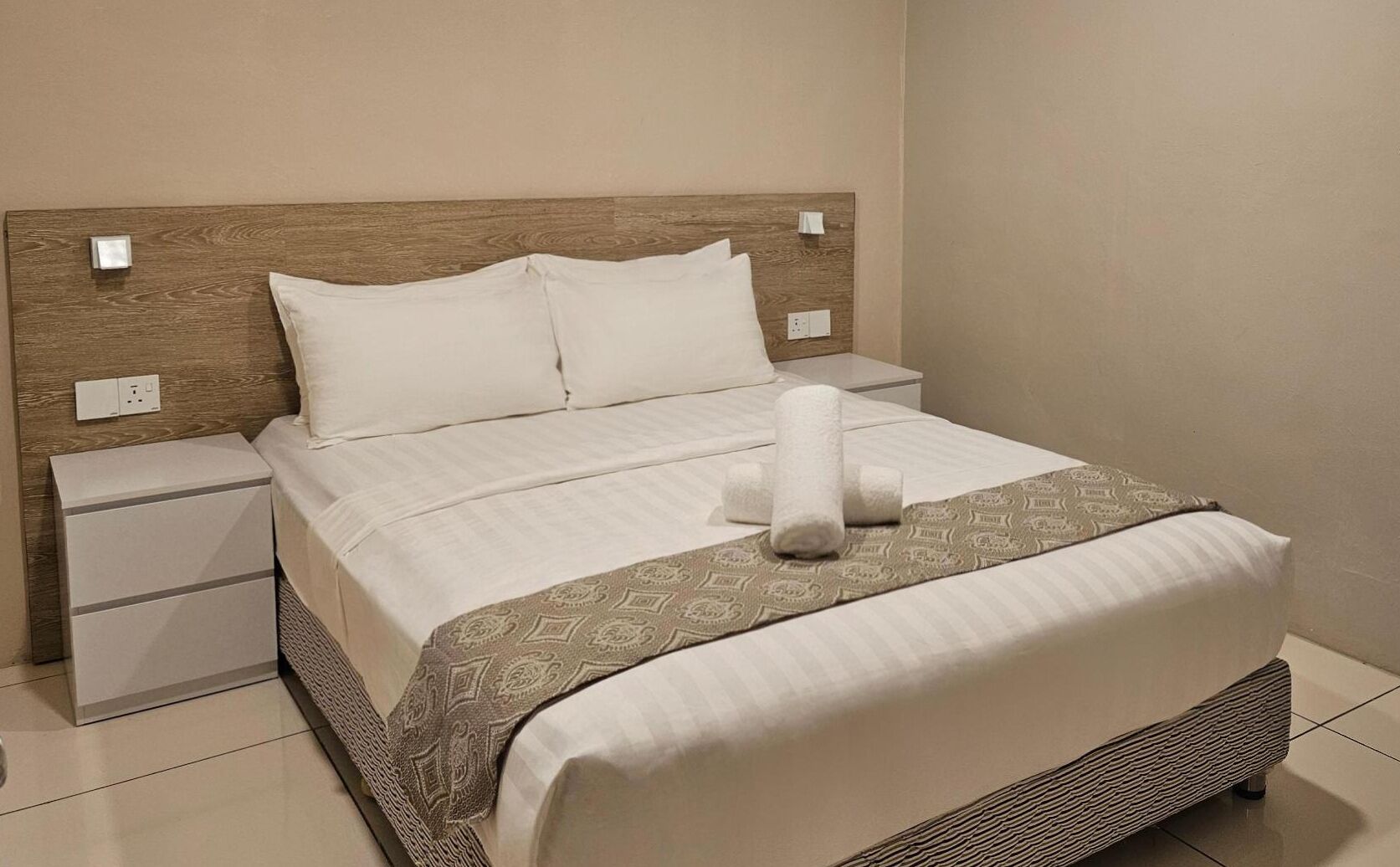 Un lit bien fait, avec du linge de lit blanc, un plaid à motifs et une serviette enroulée. On y trouve une tête de lit en bois, une table de chevet avec une lampe et une prise électrique sur le mur.
