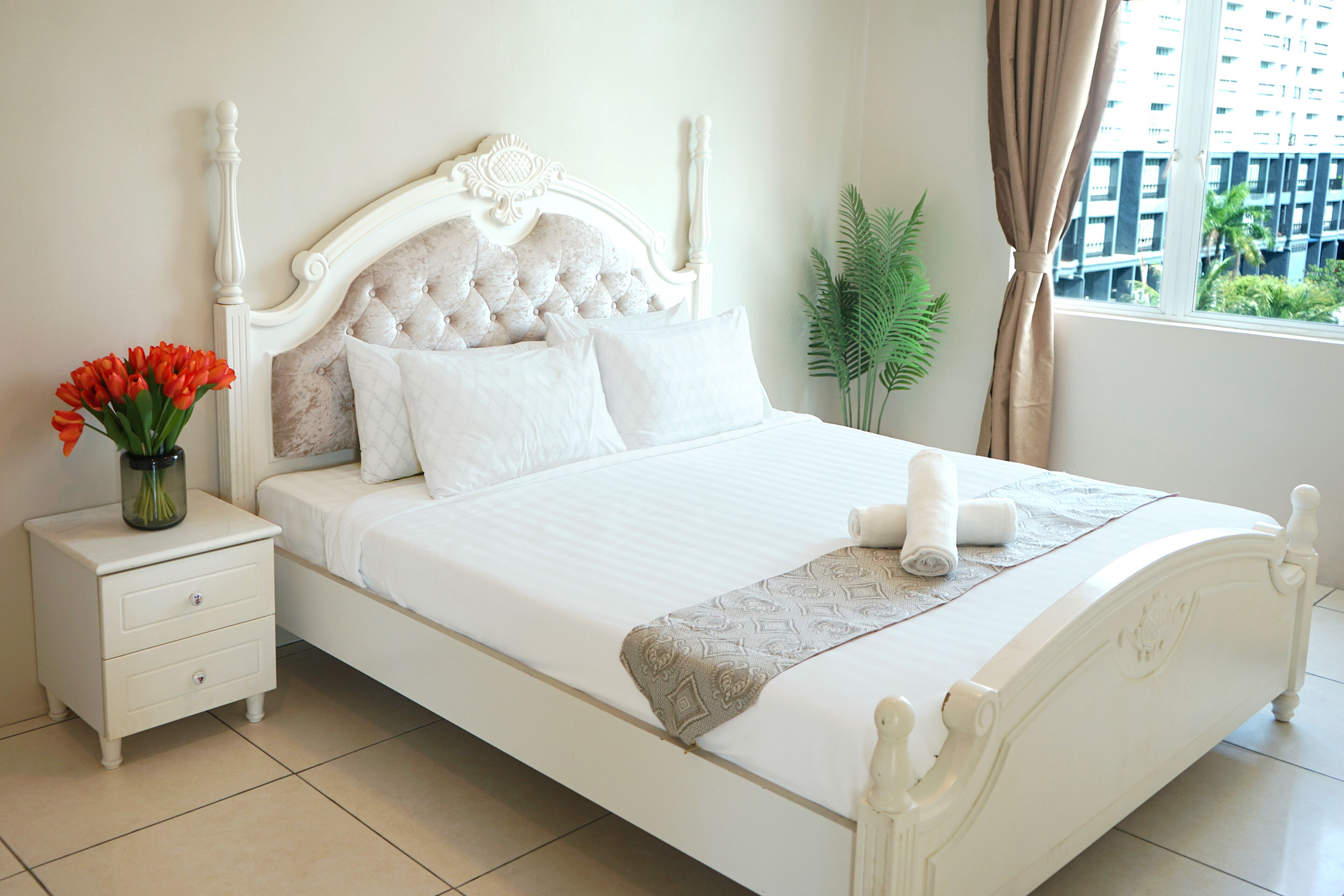 A neatly made bed with a white headboard, a bedside table with a vase of flowers, and a view of a building through a window.