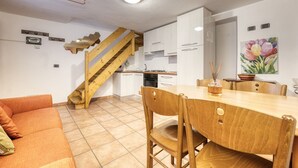 Una cucina compatta con mobili bianchi, una scala in legno, un'area pranzo con tavolo e sedie in legno, e un quadro con fiori appeso al muro.