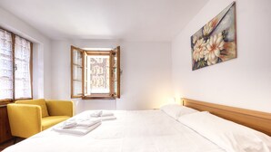 A hotel room with a bed, a yellow armchair, a window with open shutters, and a painting on the wall.