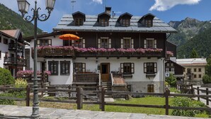 A traditional chalet with a wooden balcony, a stone entrance, and a mountain backdrop.