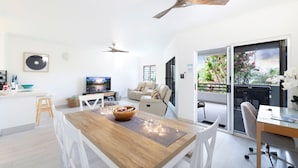 A modern living room with a wooden dining table, white chairs, a sofa, a TV, and a ceiling fan. - Nightcliff Foreshore Seaview Pool Workspace (Rapid Creek)