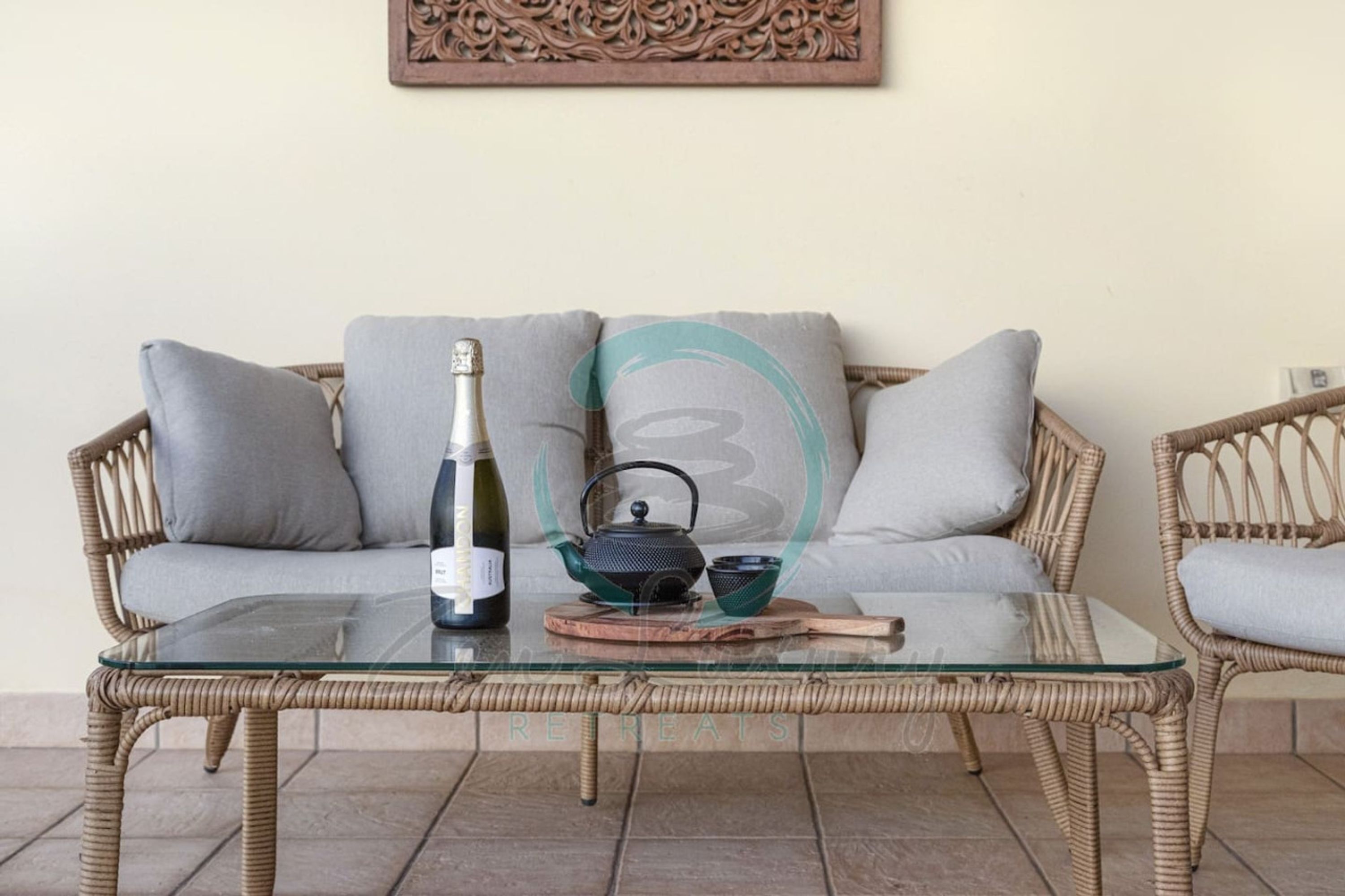 A wicker sofa with grey cushions, a glass table with a bottle, a teapot, and a cup.