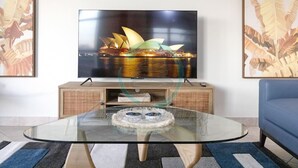 A living room with a glass coffee table, a flat-screen TV mounted on the wall, and a blue sofa. - Nightcliff Foreshore Seaview Pool Workspace (Rapid Creek)