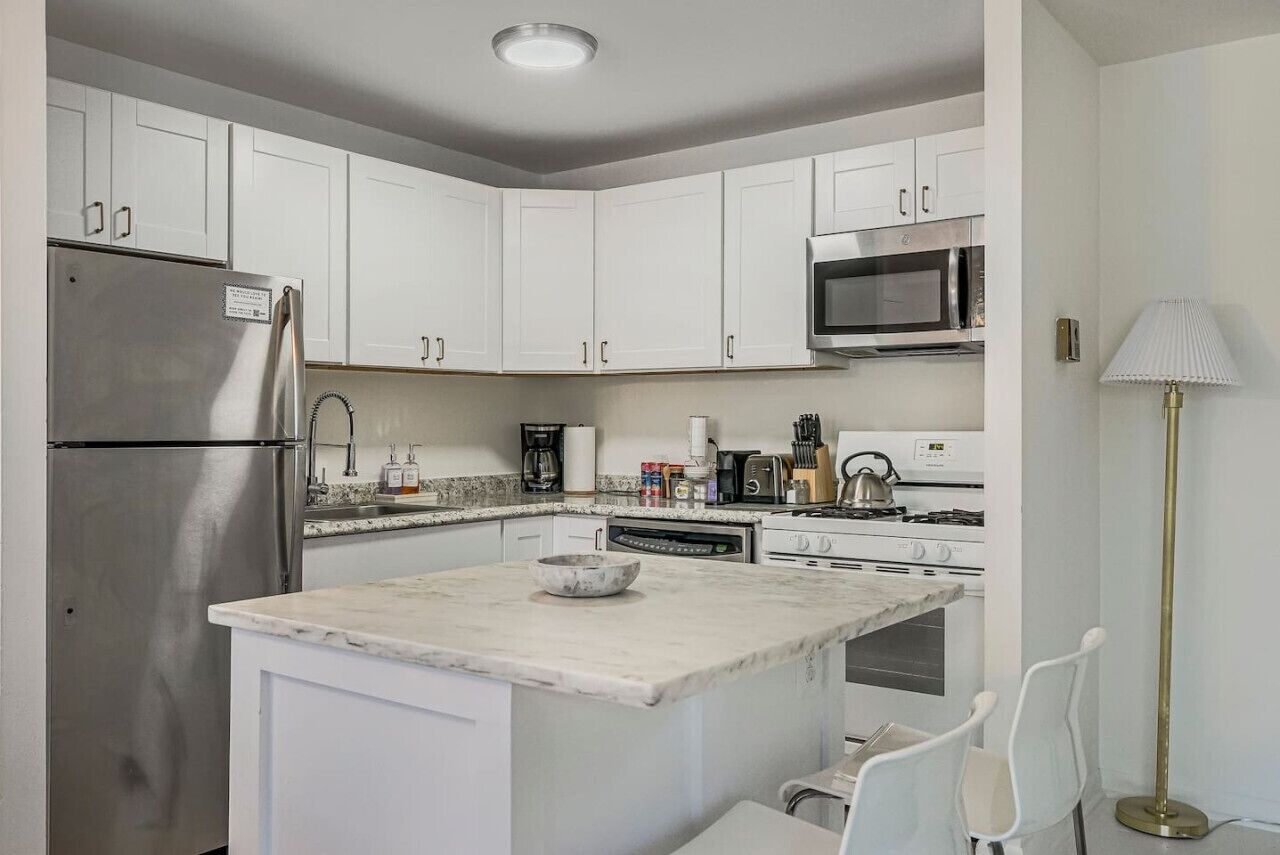 A modern kitchen with white cabinets, stainless steel appliances, a marble island, and a floor lamp.