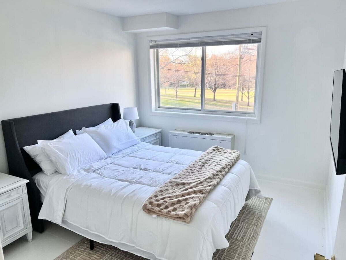 A bedroom with a bed, white bedding, a window with a view of trees, a nightstand, and a lamp.