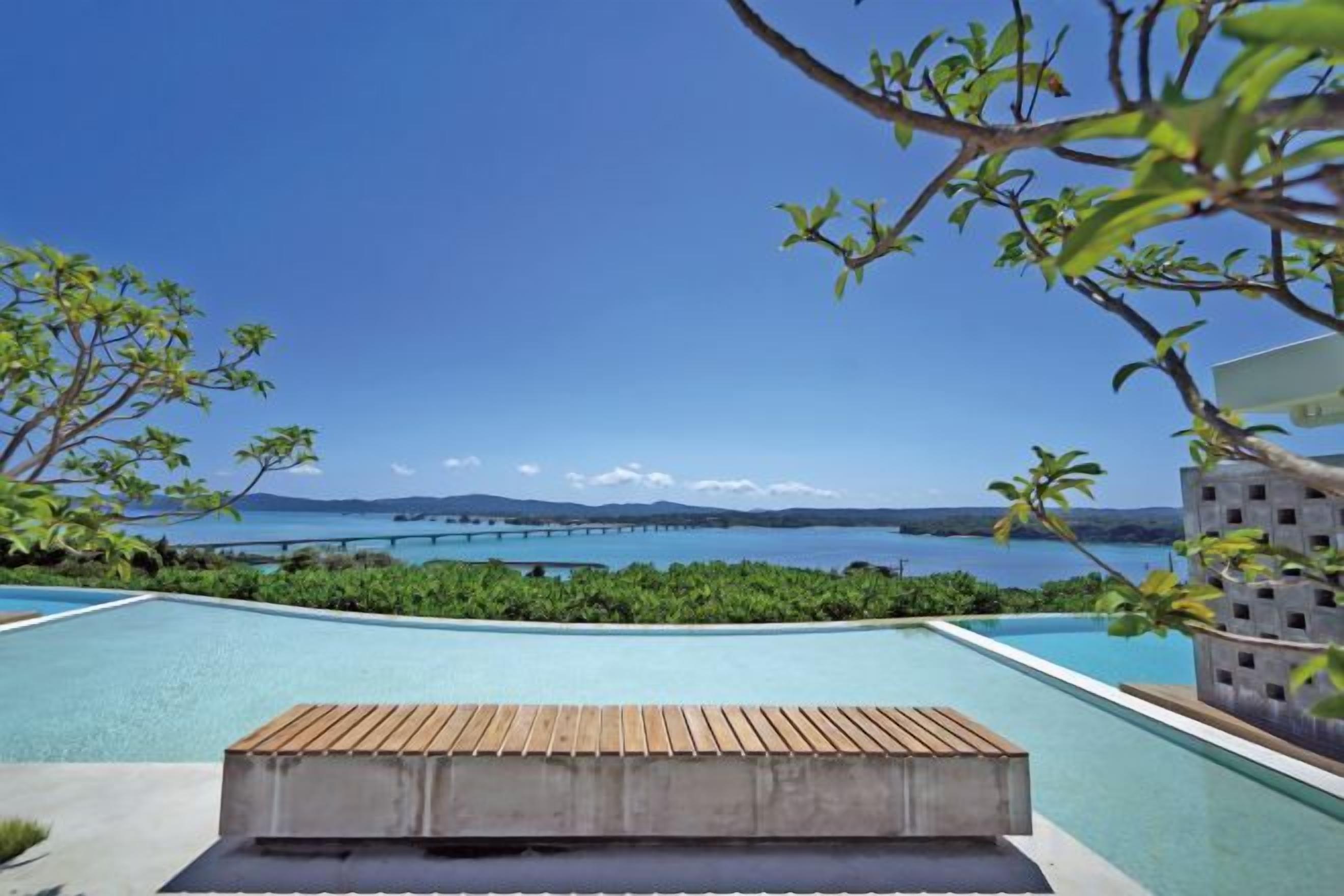 Une piscine dotée d’une terrasse en bois, entourée de verdure et offrant une vue sur une étendue d’eau avec une terre ferme visible au loin.