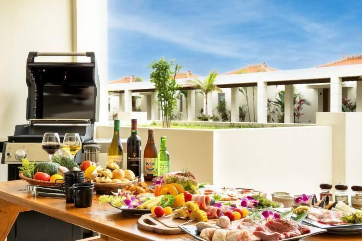 Un coin barbecue avec différentes sortes de viandes, de légumes et de condiments, un gril et une vue sur un bâtiment de style complexe hôtelier agrémenté de palmiers.