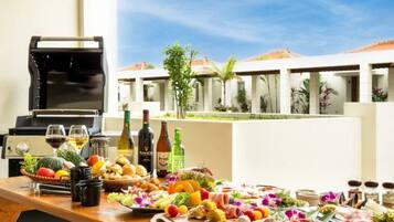 Preparación para barbacoa con diferentes carnes, verduras y salsas, una parrilla y vistas a un edificio de estilo turístico con palmeras.