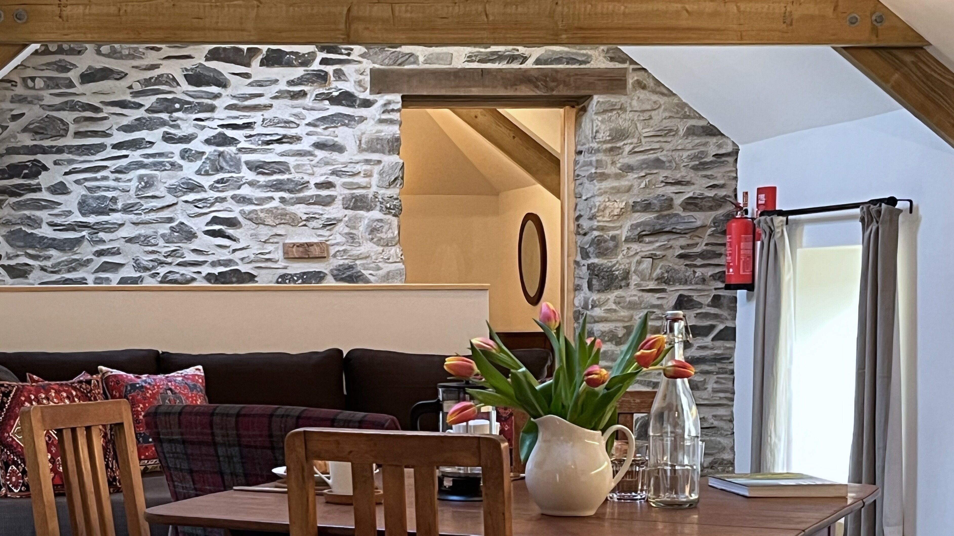 Une pièce chaleureuse avec un mur en pierre, un plafond en bois, un canapé marron, une table en bois et un vase rempli de fleurs.