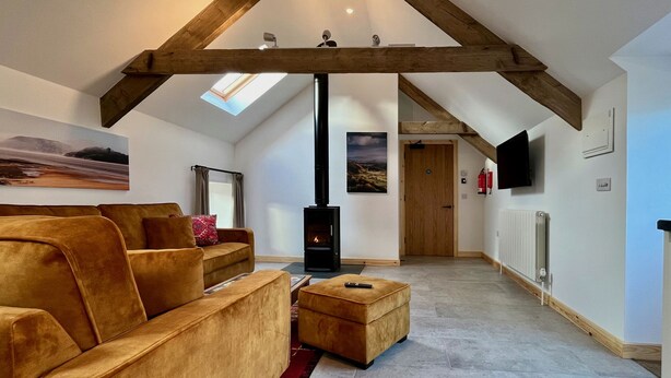 A loft-style living room with a high ceiling, exposed wooden beams, a fireplace, a sofa, a coffee table, and a television mounted on the wall. - Cambrian Cottages (Pencader)