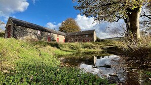 A rustic stone building with red accents, a small pond, and a tree in a natural setting. - Cambrian Cottages (Pencader)