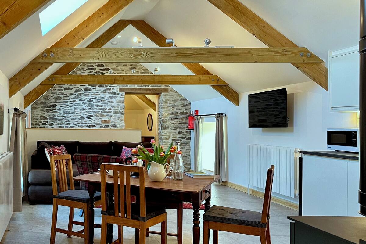A dining area with a wooden table, chairs, and a vase of tulips. There is a stone wall in the background and a television mounted on the wall.
