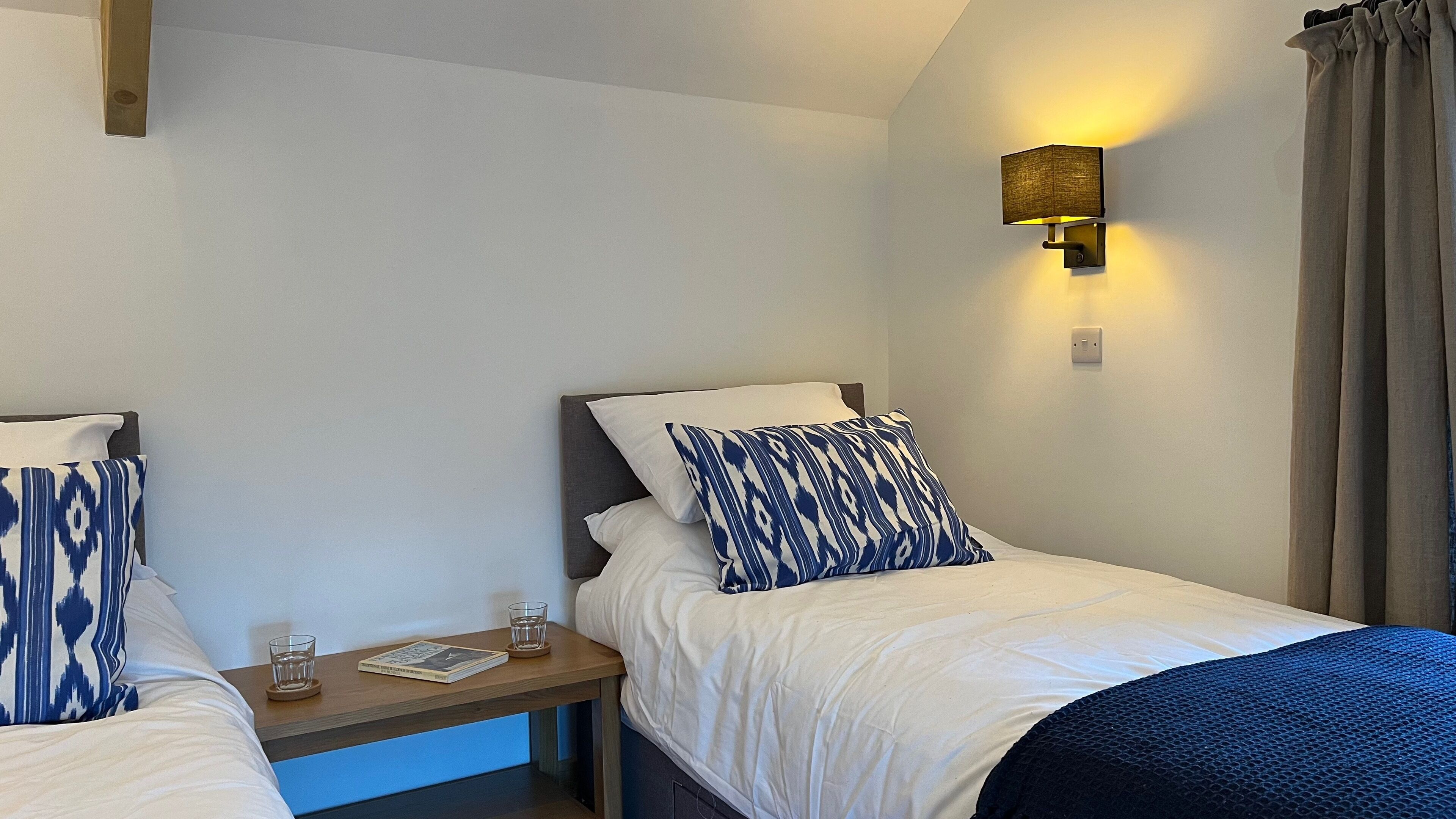 Quarto de hotel com duas camas, cada uma com almofada estampada, mesa de cabeceira com copos e um livro, e luminária fixada na parede.