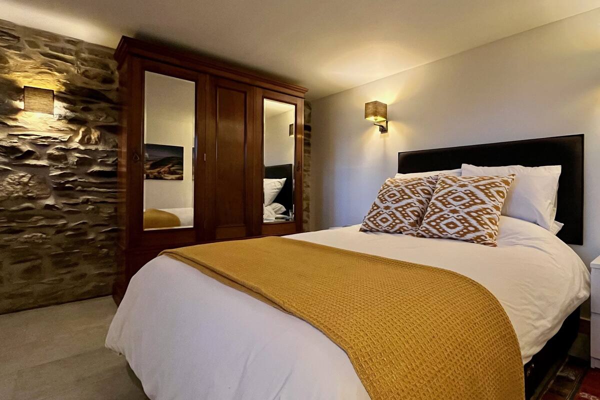 A neatly made bed with a patterned pillow and a blanket, a wooden wardrobe, a stone wall, and a bedside table with a glass and a vase.