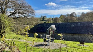 A rustic stone building with a sloped roof, a wooden fence, and a small table and chairs in the foreground. - Cambrian Cottages (Pencader)