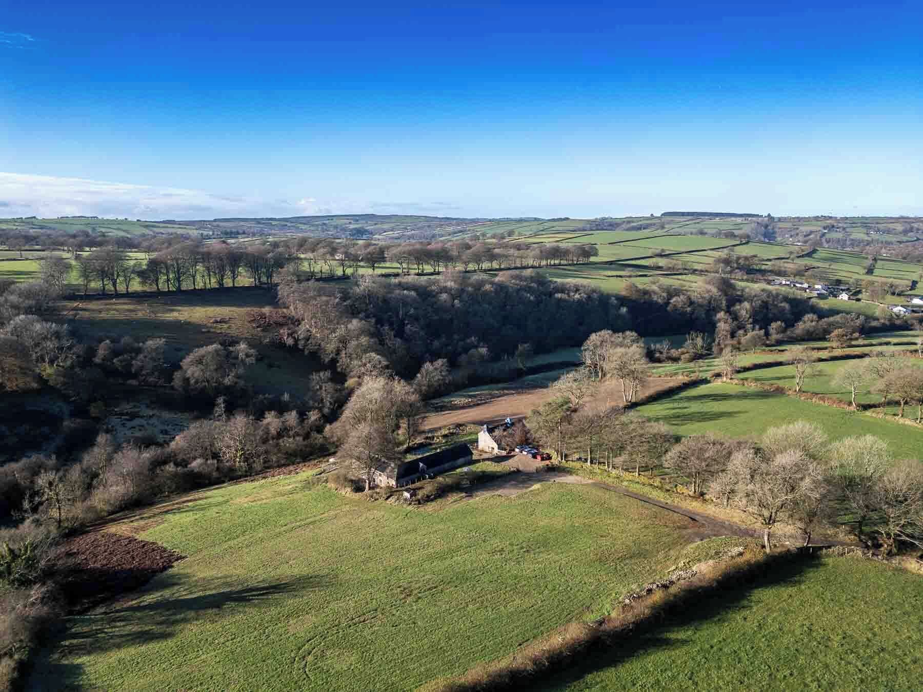 Een landelijk landschap met glooiende velden, verspreide bomen en enkele gebouwen.