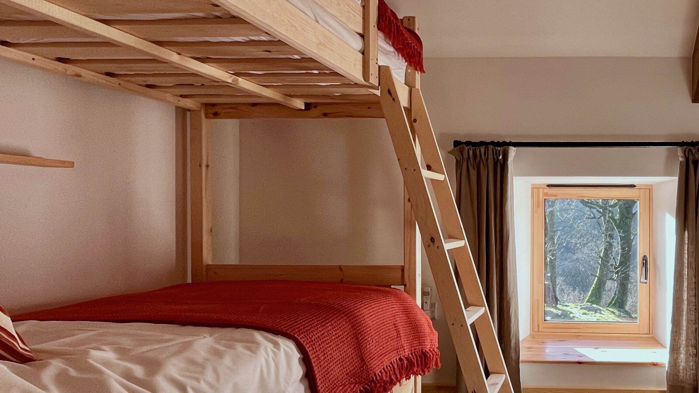 A bunk bed with a red blanket, a wooden ladder, a window with a view of trees, and a curtain.