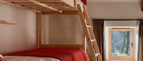 A bunk bed with a red blanket, a wooden ladder, a window with a view of trees, and a curtain.