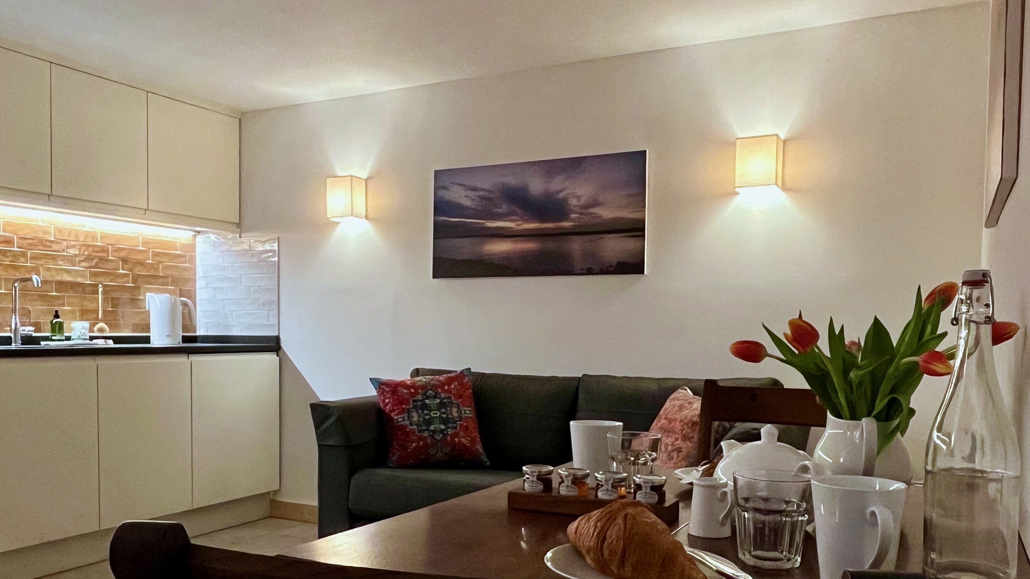 Un salon moderne avec un canapé vert, une table à manger ornée d’un vase de tulipes et un tableau représentant un coucher de soleil accroché au mur.