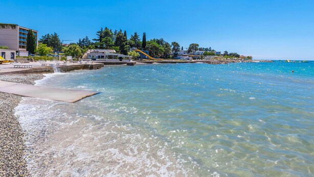 Ein Strand mit klarem Wasser, einem Rettungsturm und einem Gebäude mit der Aufschrift 'Hotel'.