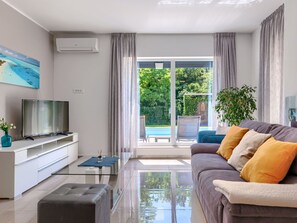A modern living room with a flat-screen TV, a glass coffee table, a sofa with cushions, and a view of a garden through large sliding glass doors.