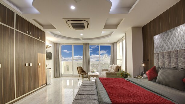 A modern bedroom with a large bed, a bench, a sofa, and a view of the sky through large windows. - SONESTA LUXURY APARTMENT (New Delhi)